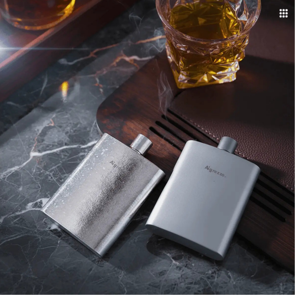 Two Aigrezzo titanium hip flasks beside a glass of whiskey, minimalist lightweight titanium flask for refined everyday carry