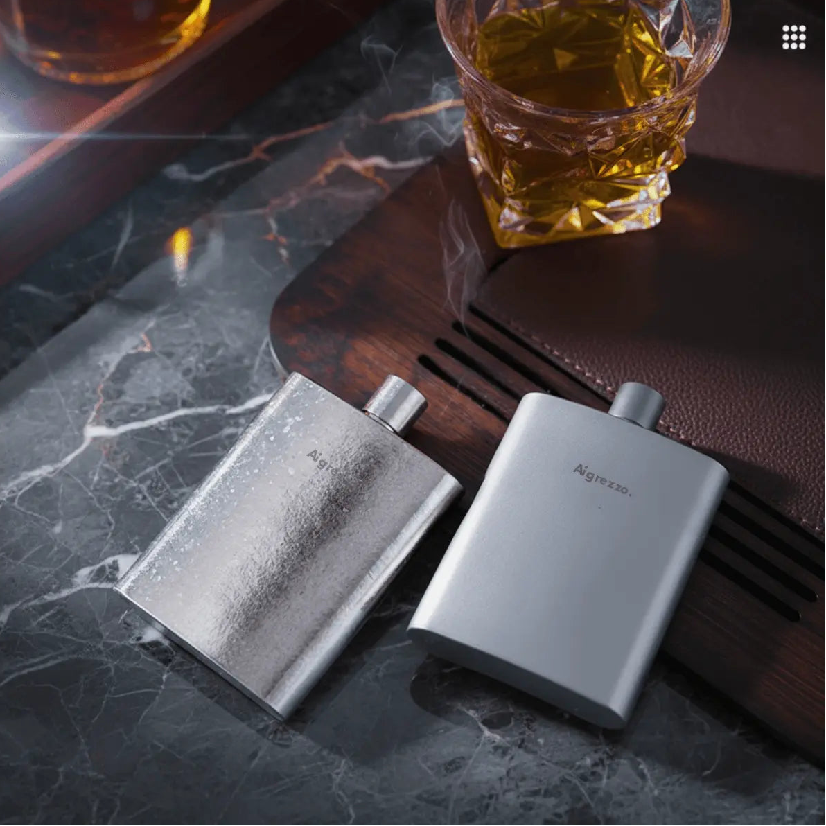 Two Aigrezzo titanium hip flasks beside a glass of whiskey, minimalist lightweight titanium flask for refined everyday carry